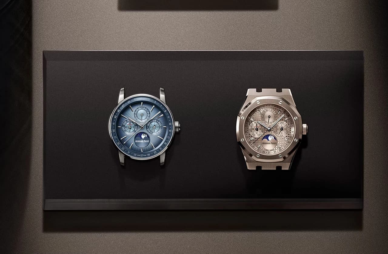dos relojes Audemars Piguet con calendario perpetuo y esfera lunar, uno redondo de esfera azul y otro octagonal de esfera plateada, expuestos sobre una base negra