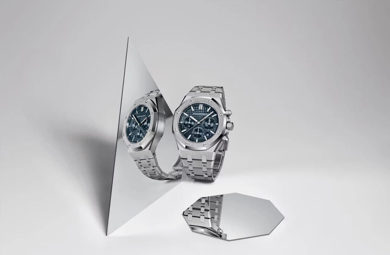 reloj de acero con esfera azul y bisel octagonal reflejado en un espejo triangular sobre fondo gris claro