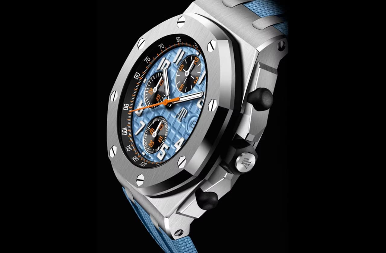 reloj cronógrafo con bisel octogonal de acero, esfera azul texturizada con tres contadores negros y detalles naranjas, correa azul, visto en ángulo