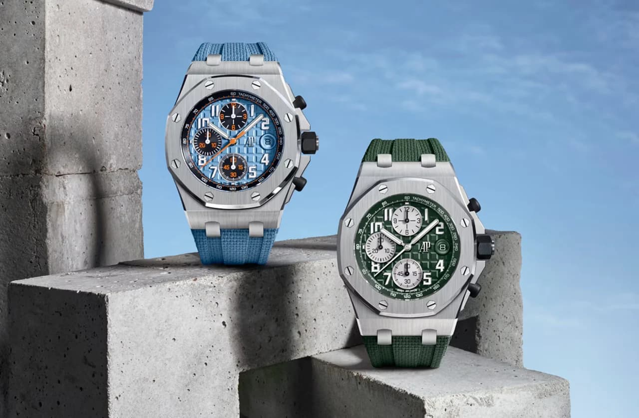dos relojes cronógrafo de caja octogonal en acero, uno con esfera y correa azul claro y otro con esfera y correa verde, apoyados sobre bloques de hormigón contra un cielo azul